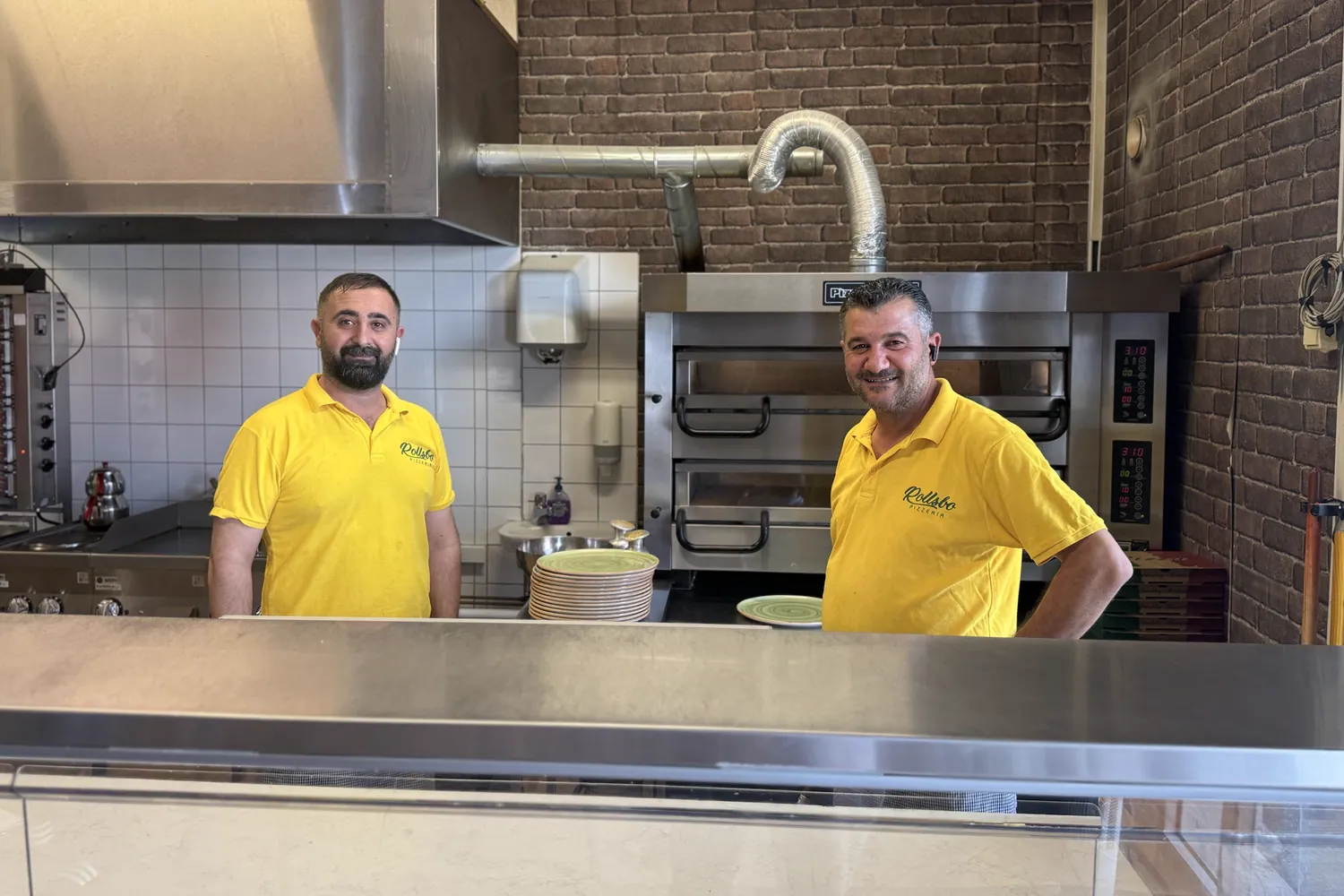 Två ur personalen på Rollsbo Pizzeria i gula tröjor bakom disken, med kebabgrillen och pizzaugnen bakom sig